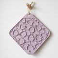A lavender silicone hanging mat with a repeating pattern of small cartoon faces, suspended by a wooden dowel and string against a plain white wall. The brand name 'somaso' is printed at the bottom.