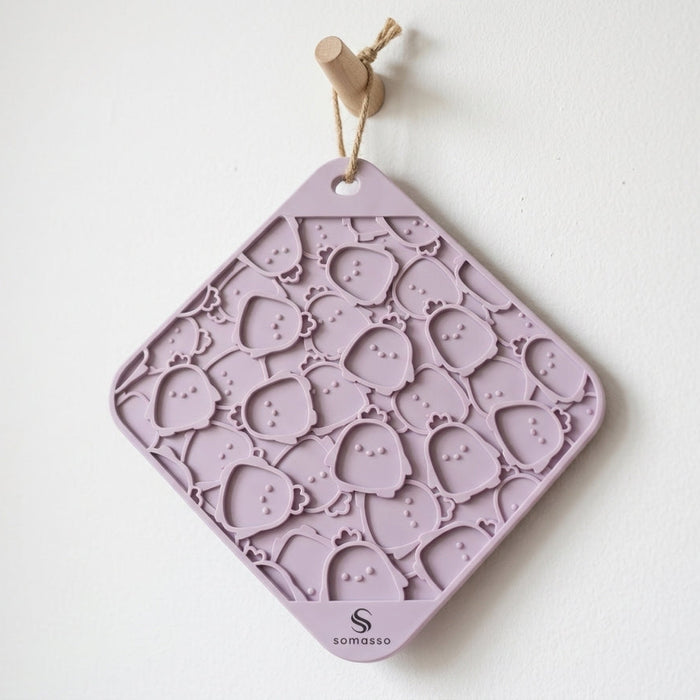 A lavender silicone hanging mat with a repeating pattern of small cartoon faces, suspended by a wooden dowel and string against a plain white wall. The brand name 'somaso' is printed at the bottom.
