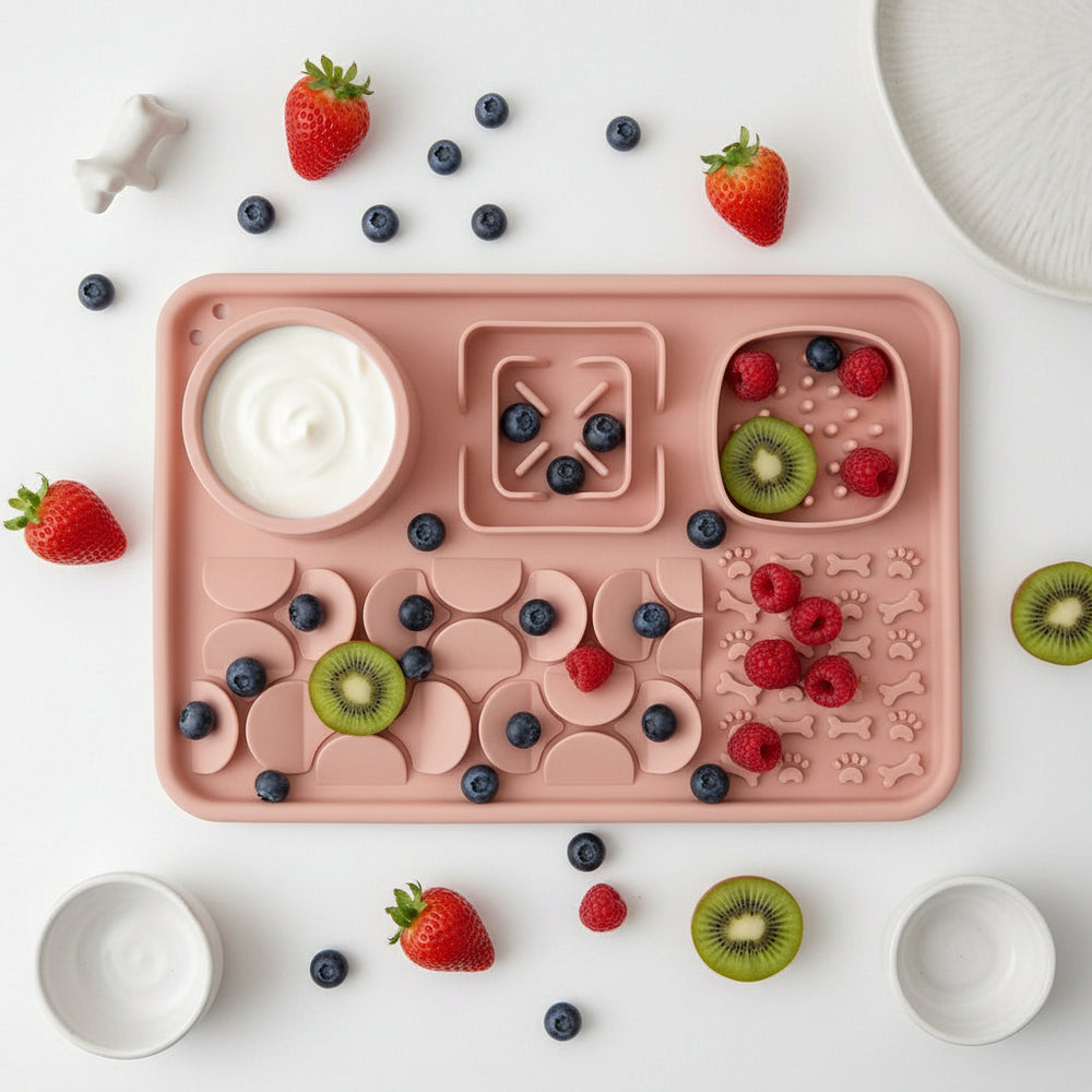 A pink silicone baby feeding tray with multiple compartments, including a round bowl with the 'Sofrito' brand logo, arranged on a white surface with small white ceramic dishes and a small white figurine.