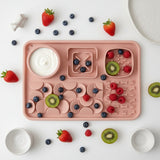 A pink silicone baby feeding tray with multiple compartments, including a round bowl with the 'Sofrito' brand logo, arranged on a white surface with small white ceramic dishes and a small white figurine.