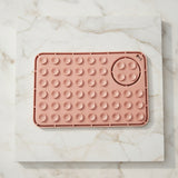 A rectangular pink silicone mat with multiple raised circular suction cups and a circular opening in the lower right, shown against a plain light background.